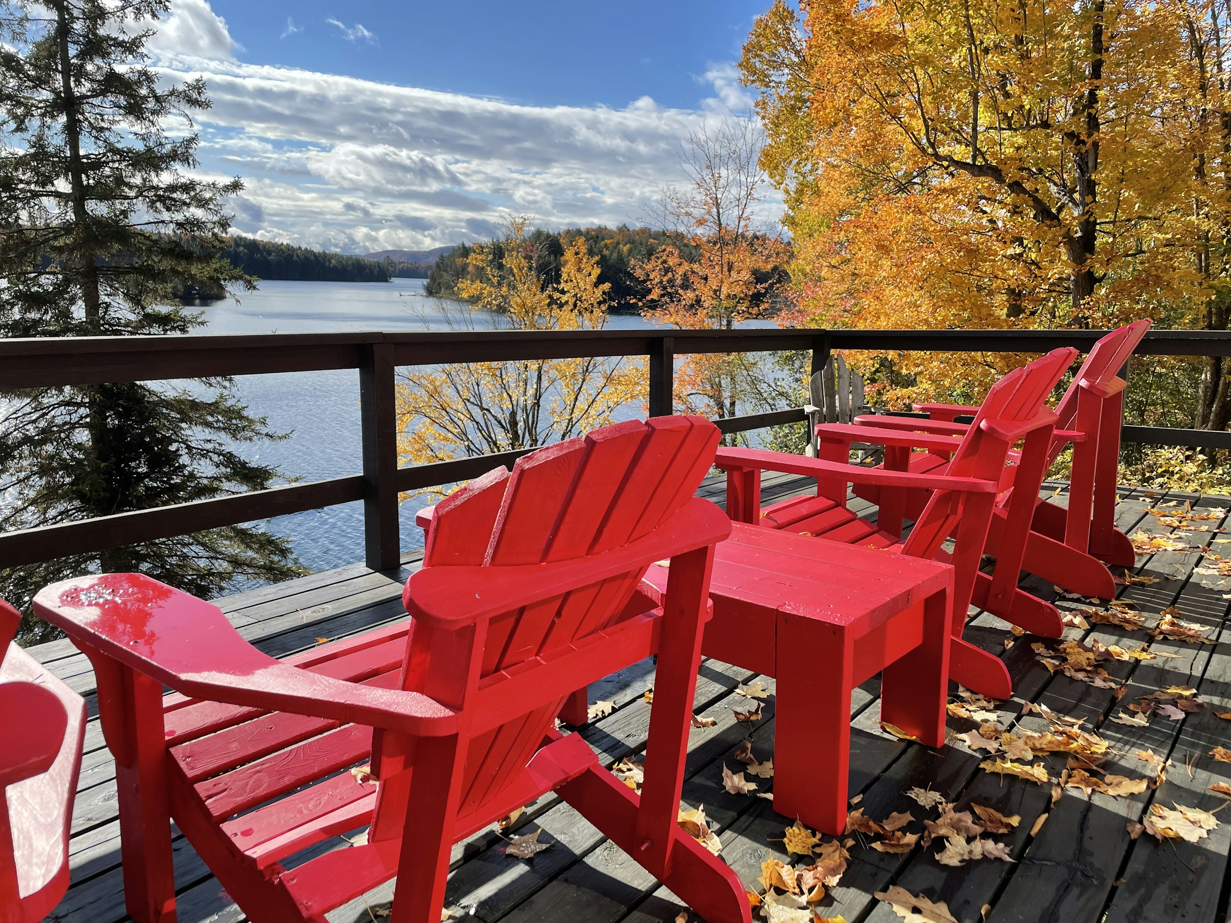 Two red Adirondack chairs on a wooden lakeside deck with autumn maples and a calm lake behind