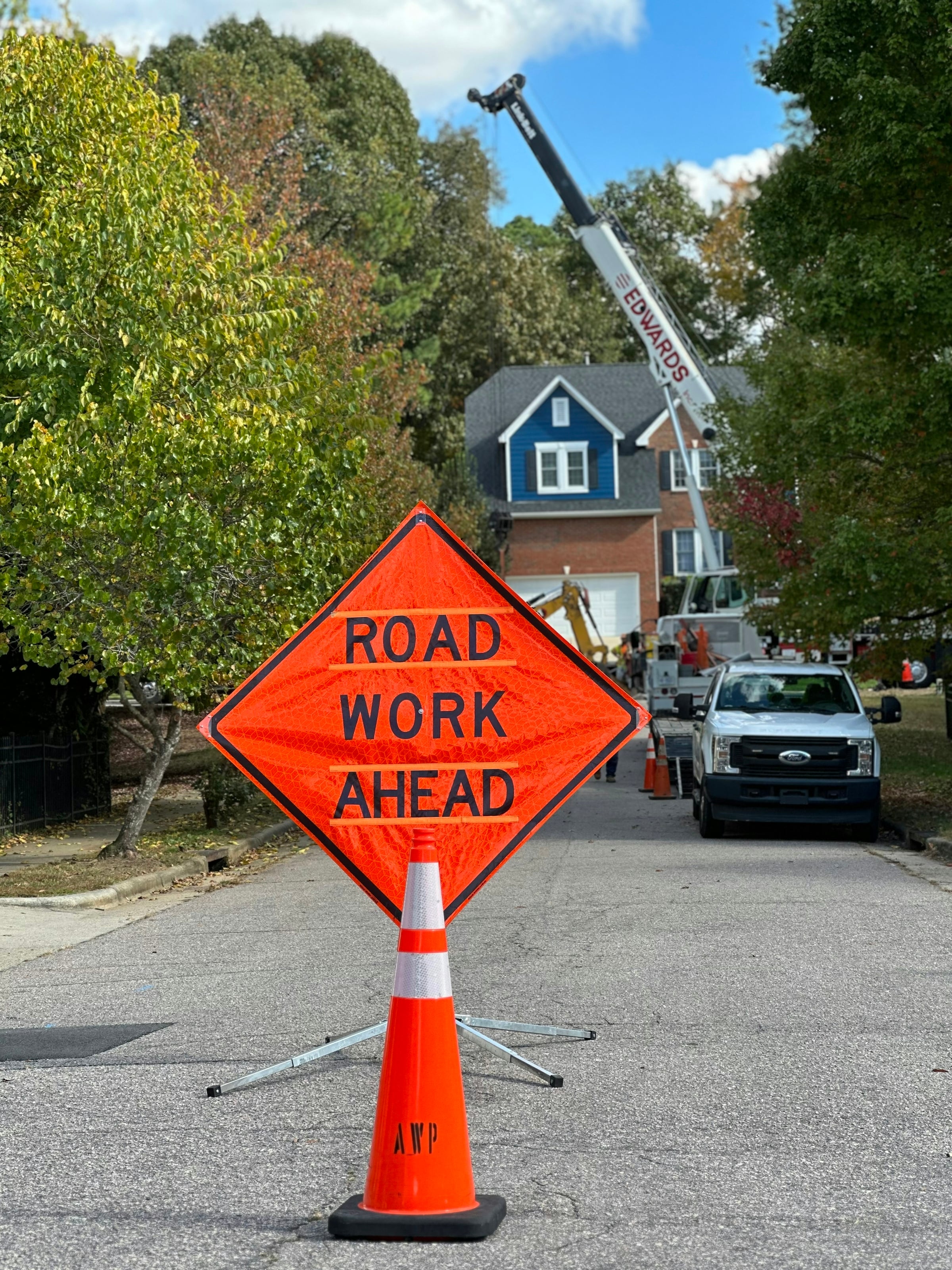 Orange Road Work Ahead sign on a traffic cone in the middle of a residential street, with a utility truck and a blue house behind it