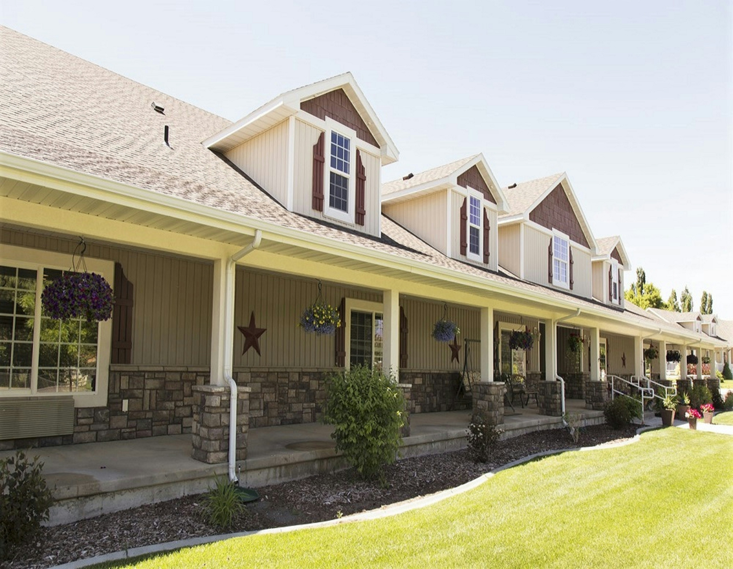 Low-rise senior living residence with a long covered front porch, stone-base columns, dormer windows and a green lawn
