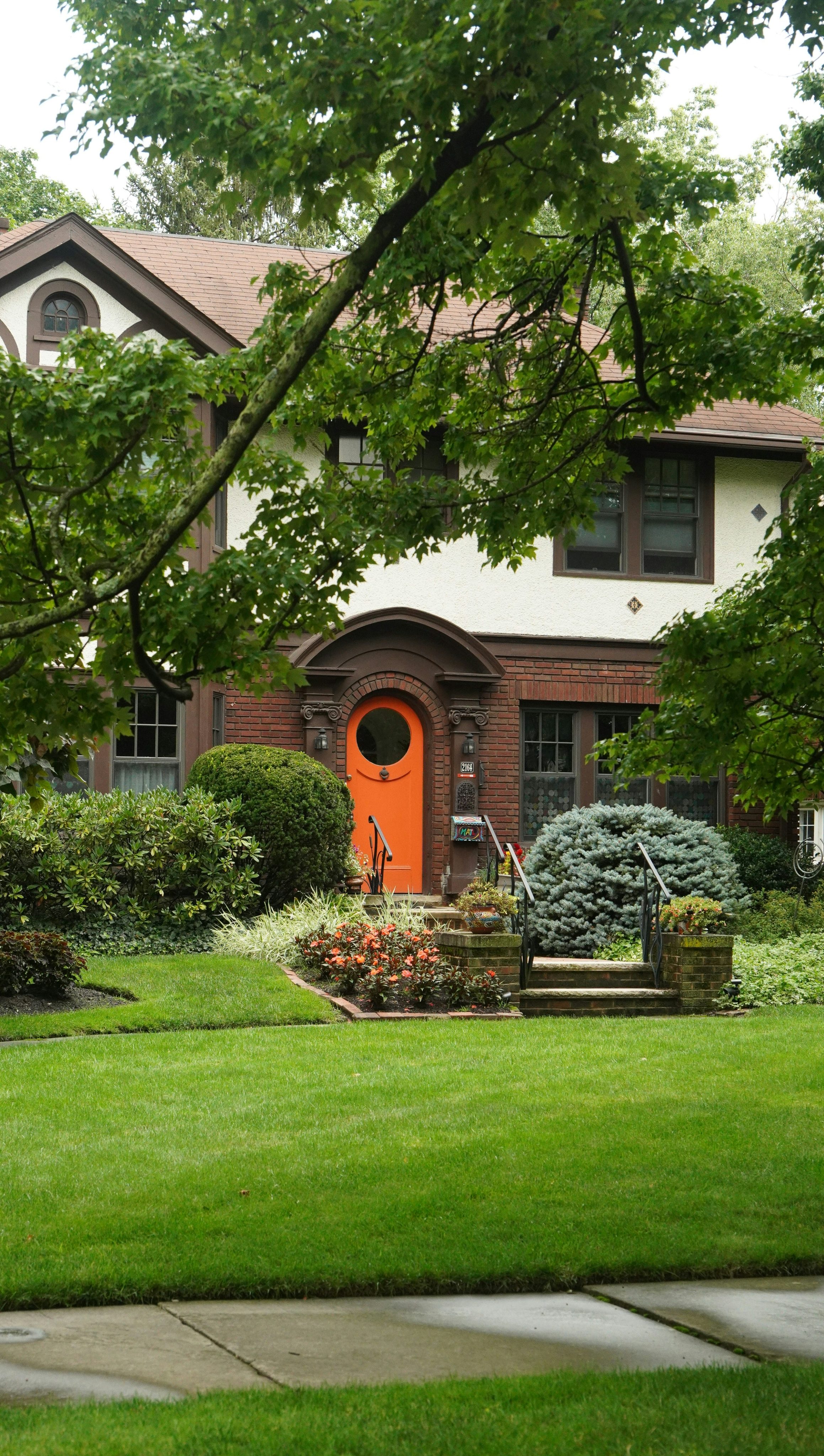Tudor-style house with an orange arched front door, rounded shrubs and a green front lawn beside a sidewalk