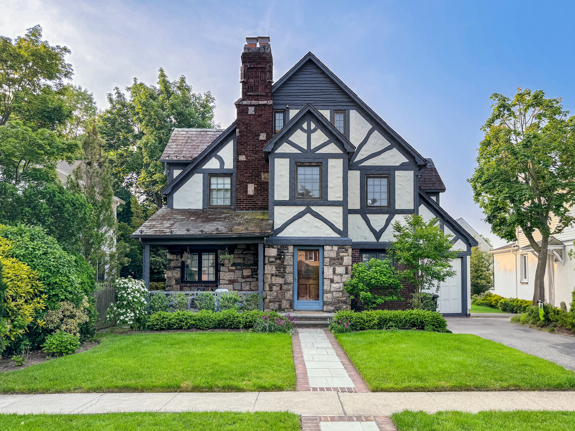 A two-story Tudor-style house with a stone chimney, dark timber trim and a landscaped front lawn