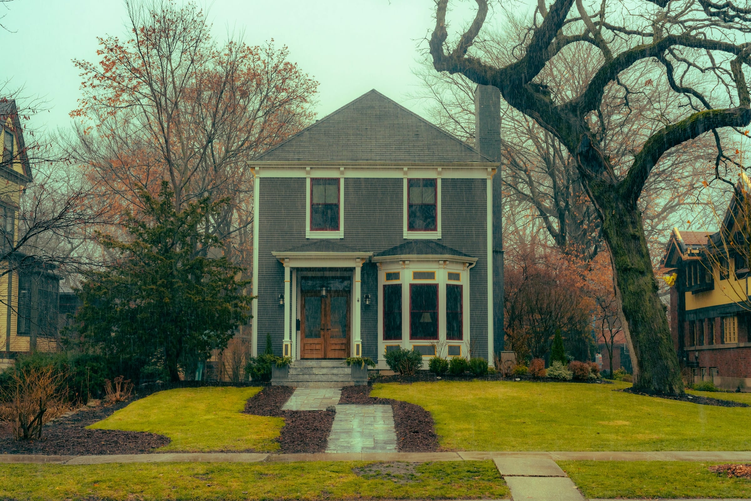 Two-story gray-green house with a front porch, a walkway and a bare tree in the front yard