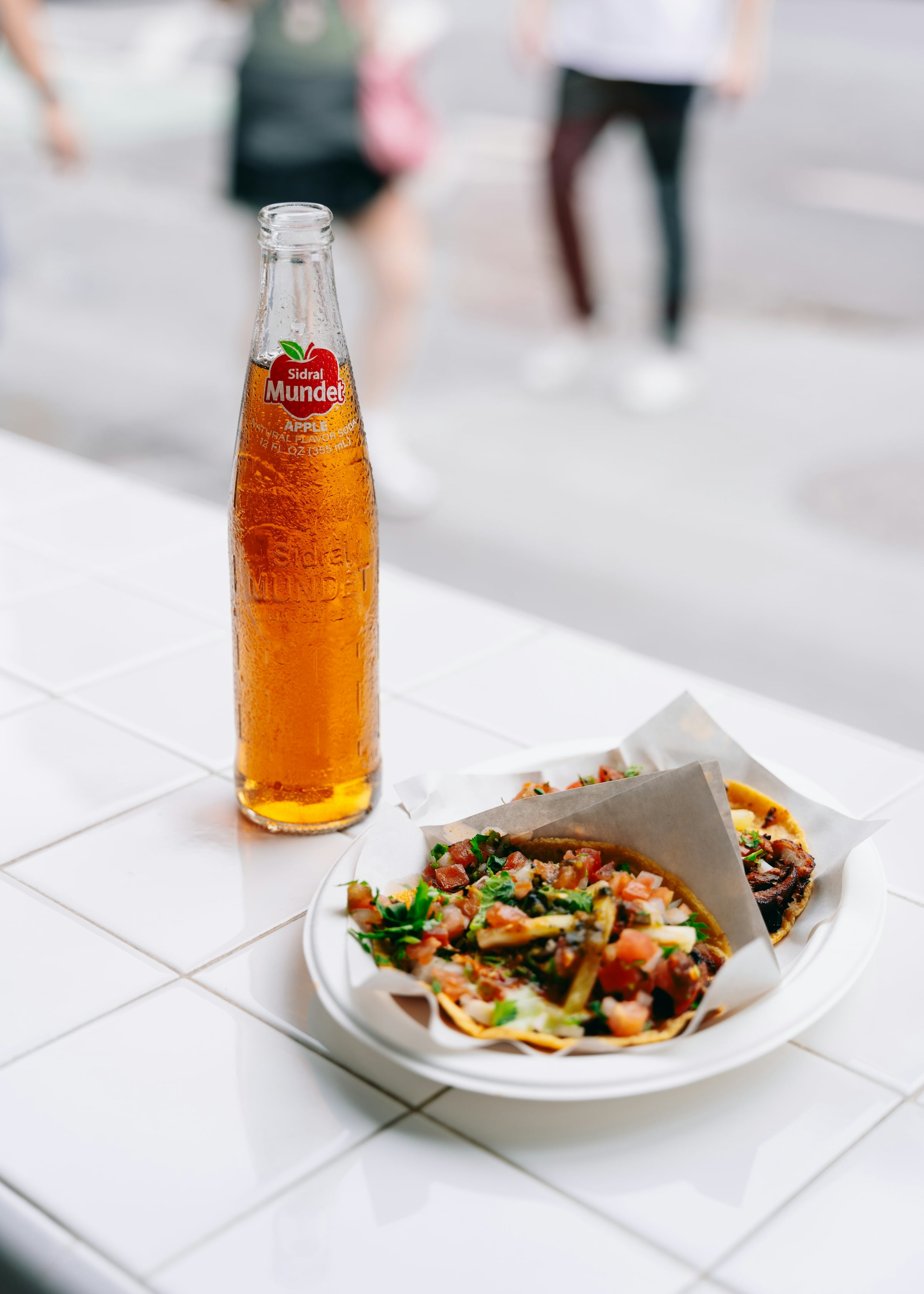 Tacos served on a paper-lined plate beside a glass soda bottle on a white tiled table, with pedestrians blurred in the background