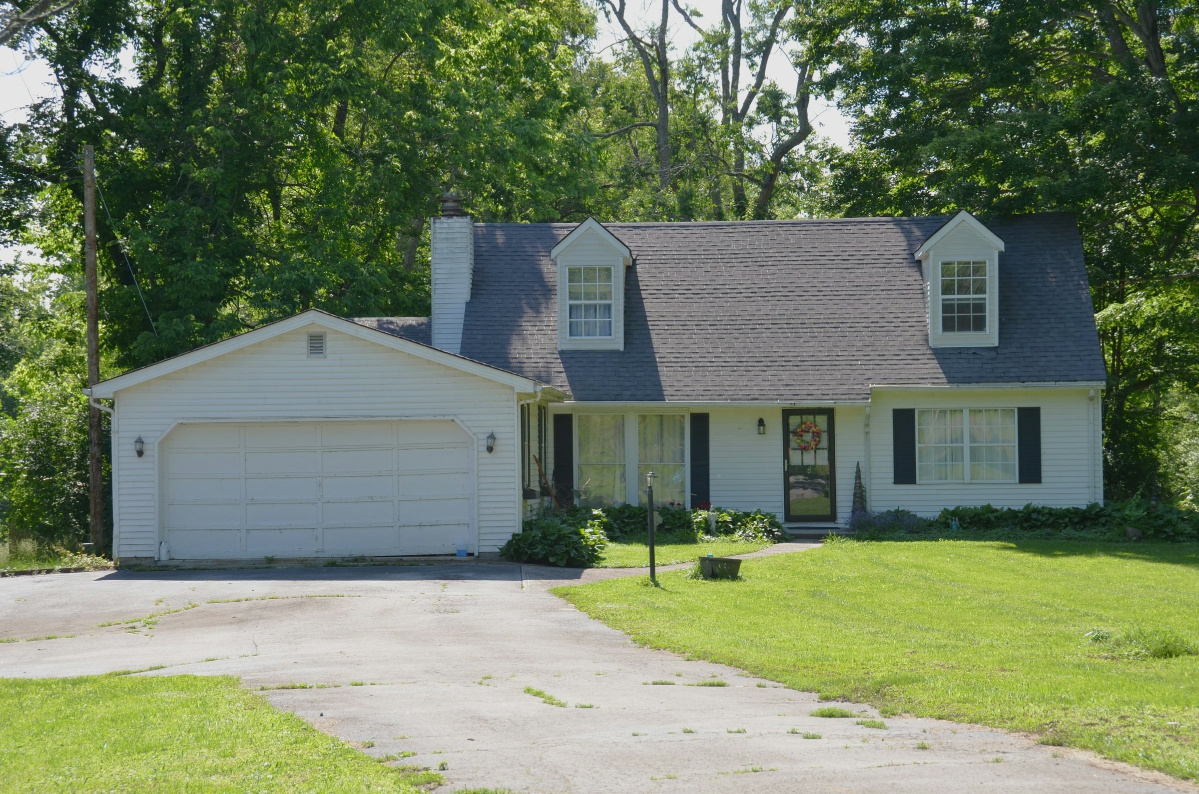 White Cape-style house with two dormers, an attached garage, a wide driveway and a green lawn under mature trees