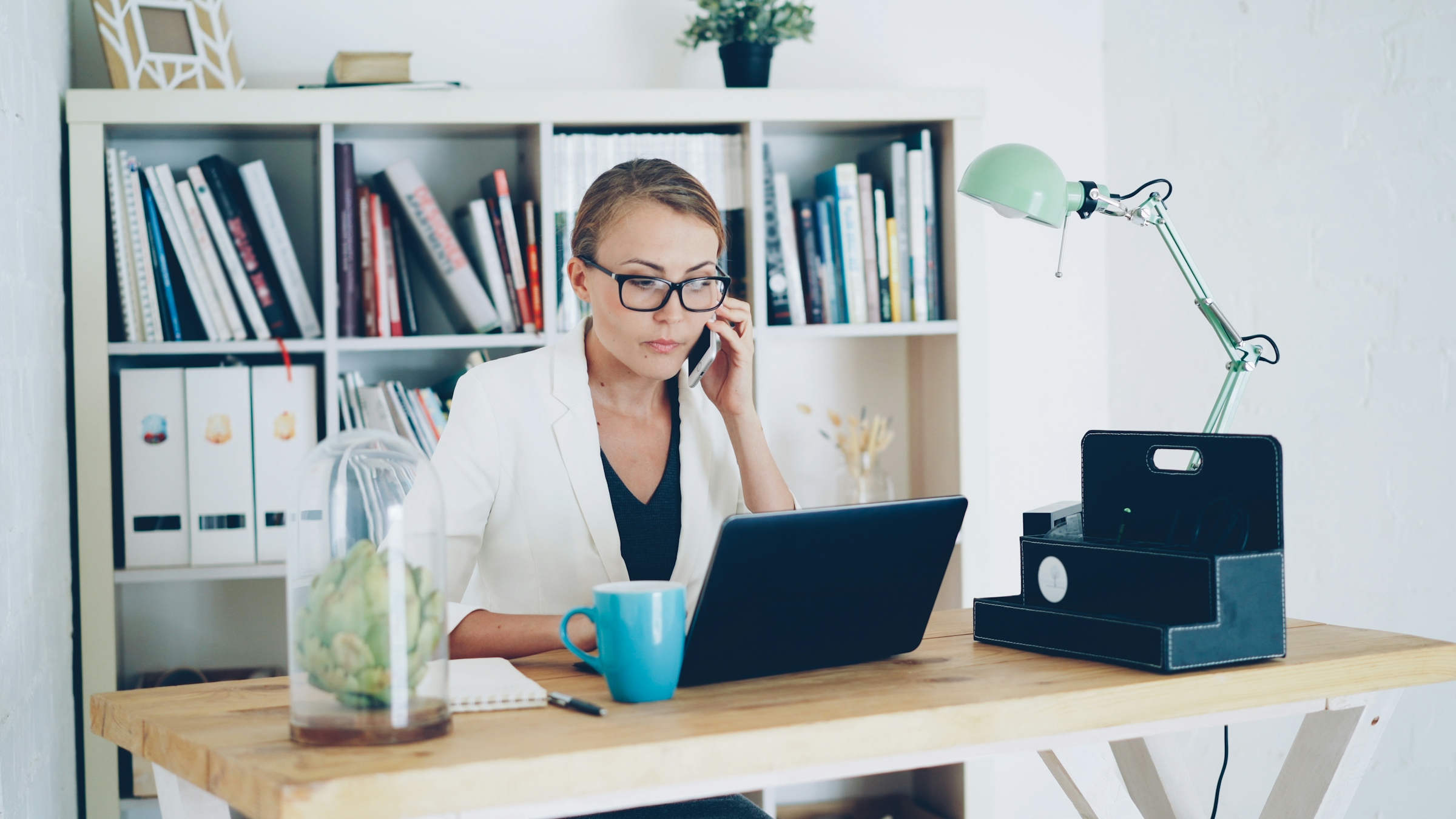 Woman with glasses on a phone call at a laptop in front of a bookshelf