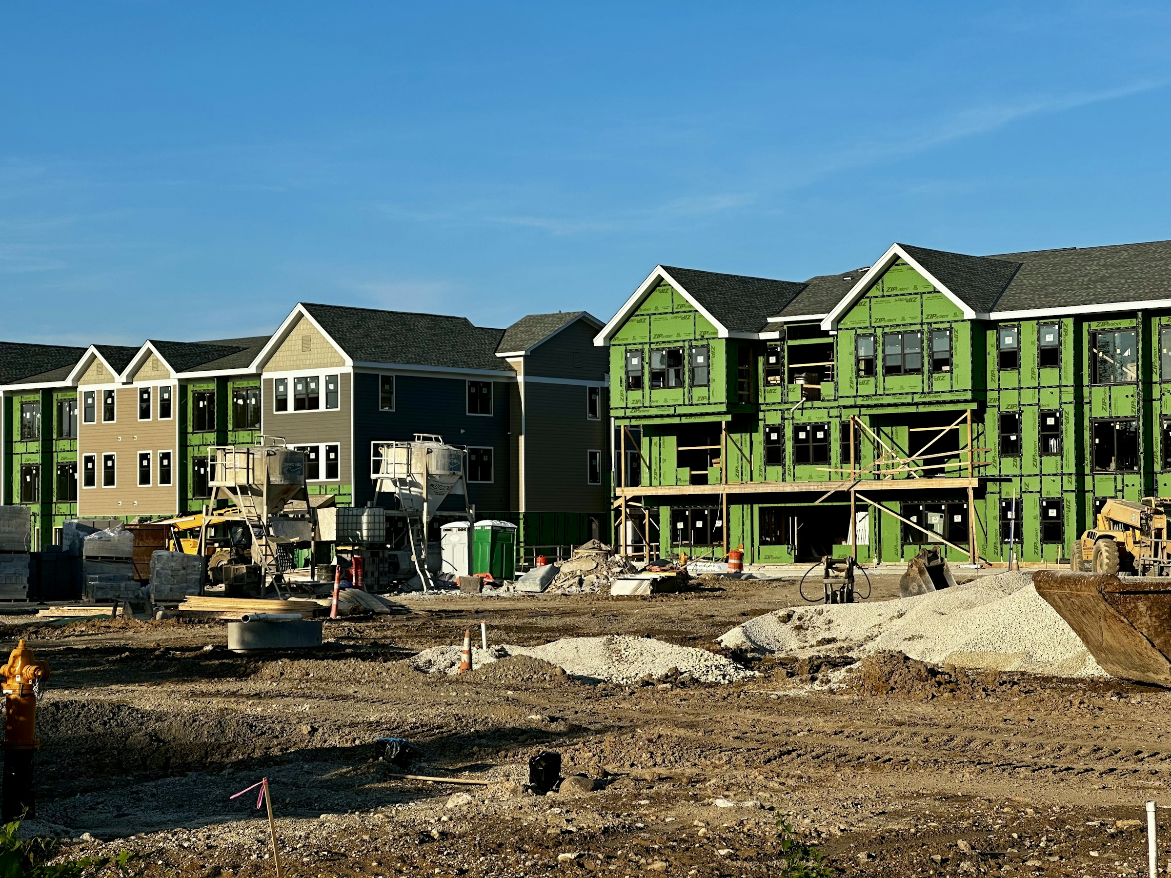 Rows of three-story wood-frame apartment buildings under construction with green sheathing and an excavator in the foreground