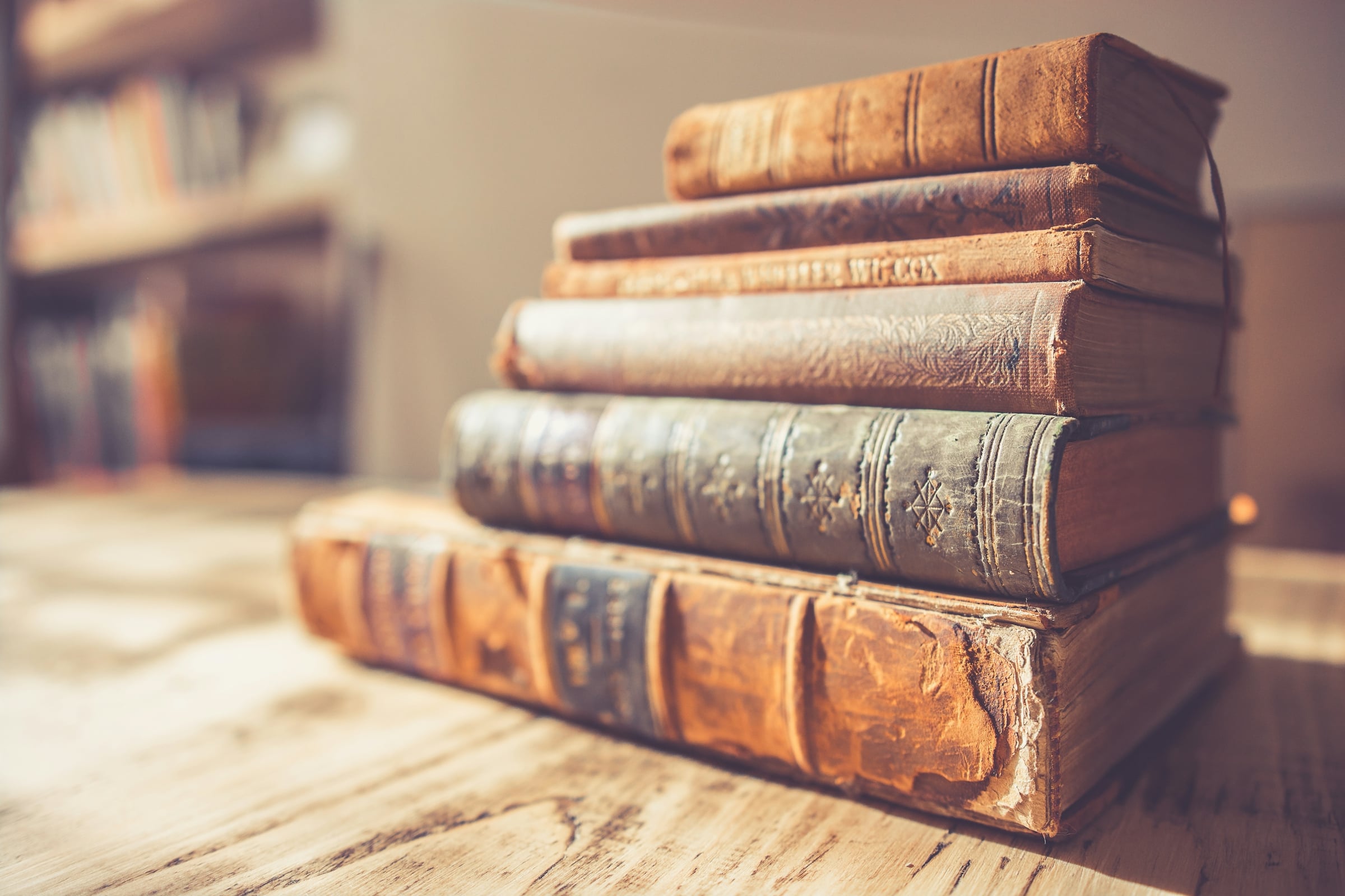 Stack of five worn leather-bound volumes on a wooden table beside a bookshelf in warm light