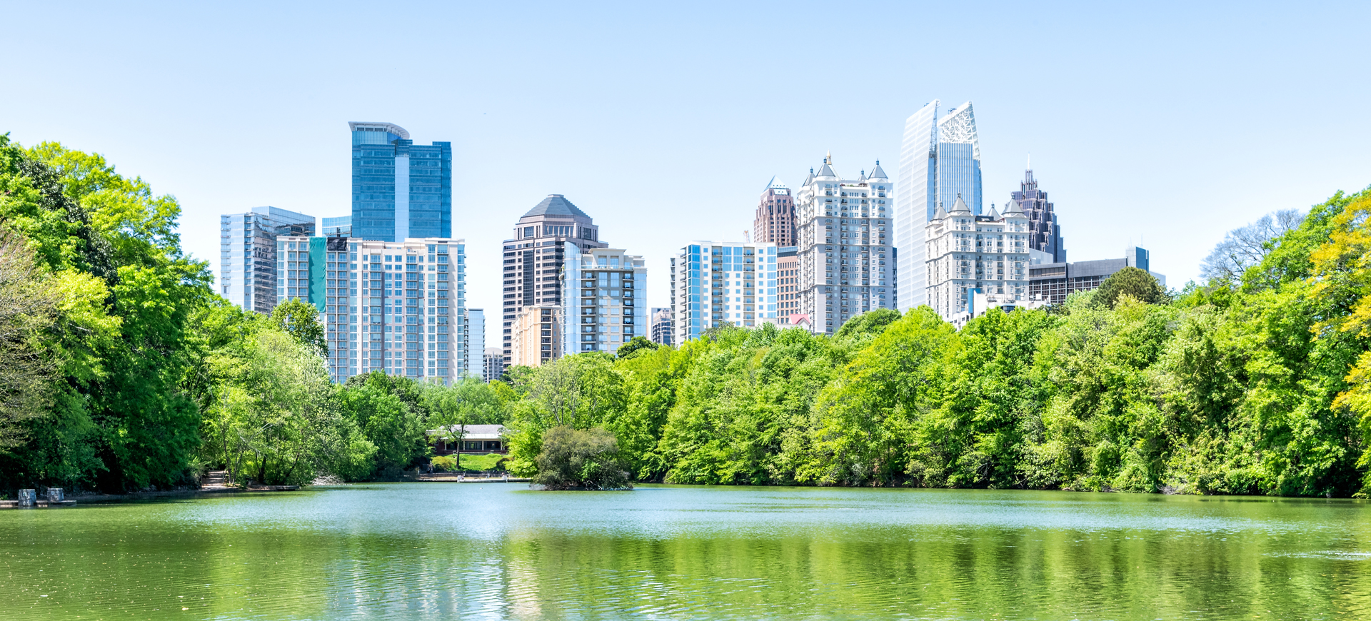 Cityscape,-skyline-view-in-Piedmont-Park-in-Atlanta,-Georgia-green-foliage,-trees,-scenic-water,-urban-city-skyscrapers-downtown-at-Lake-Clara-Meer-by-railing