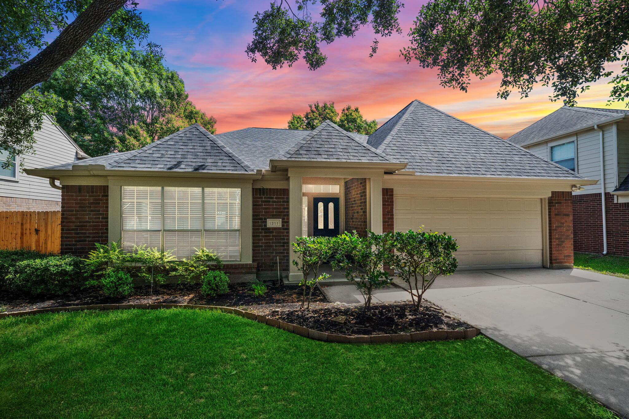 Front exterior of a single-story brick home with attached two-car garage, landscaped yard, and sunset sky