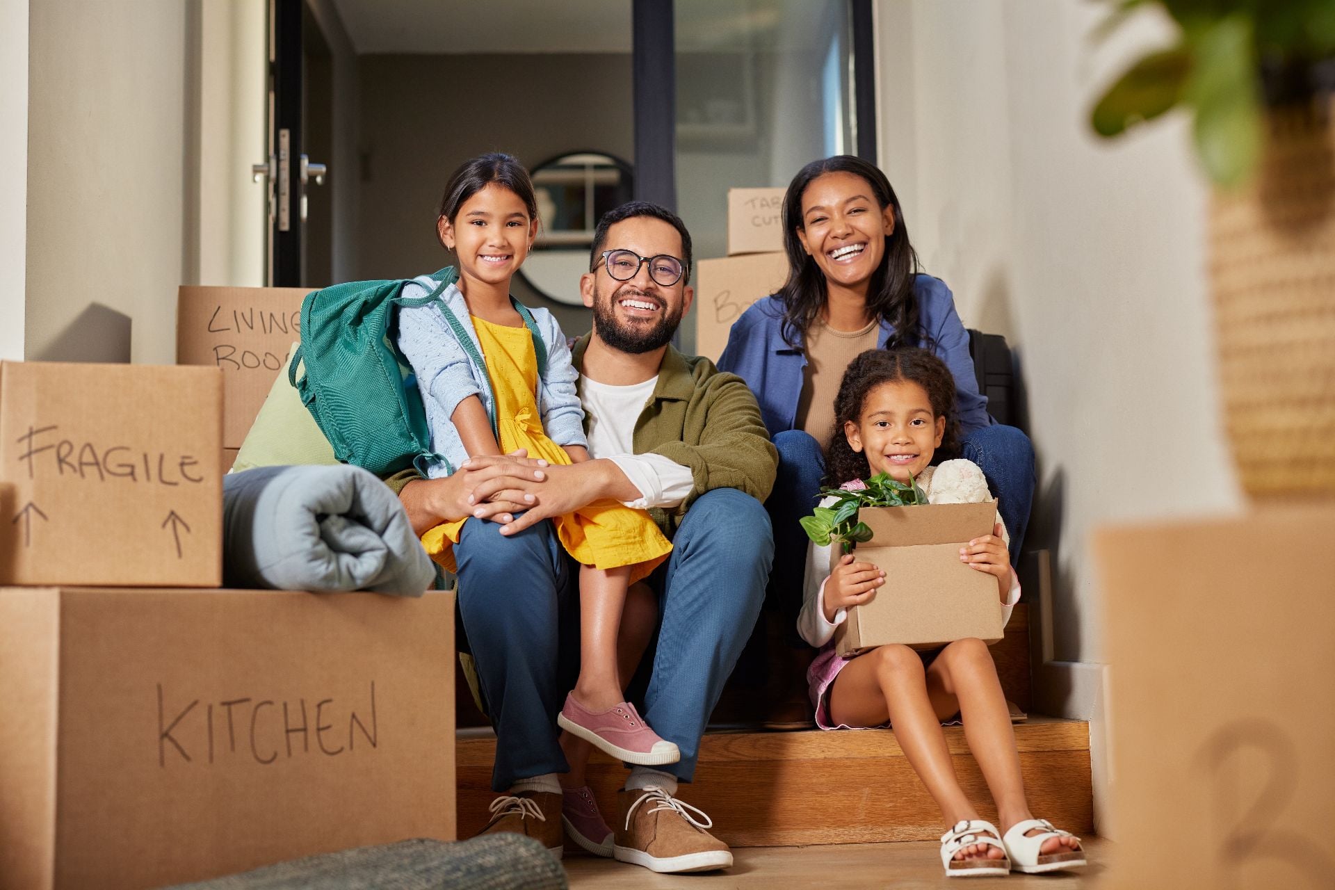 Middle eastern family sitting on stairs surrounded by moving boxes. Indian parents and their two daughters smiling on moving day. Mixed race family with daughters enjoy a moment of relax together
