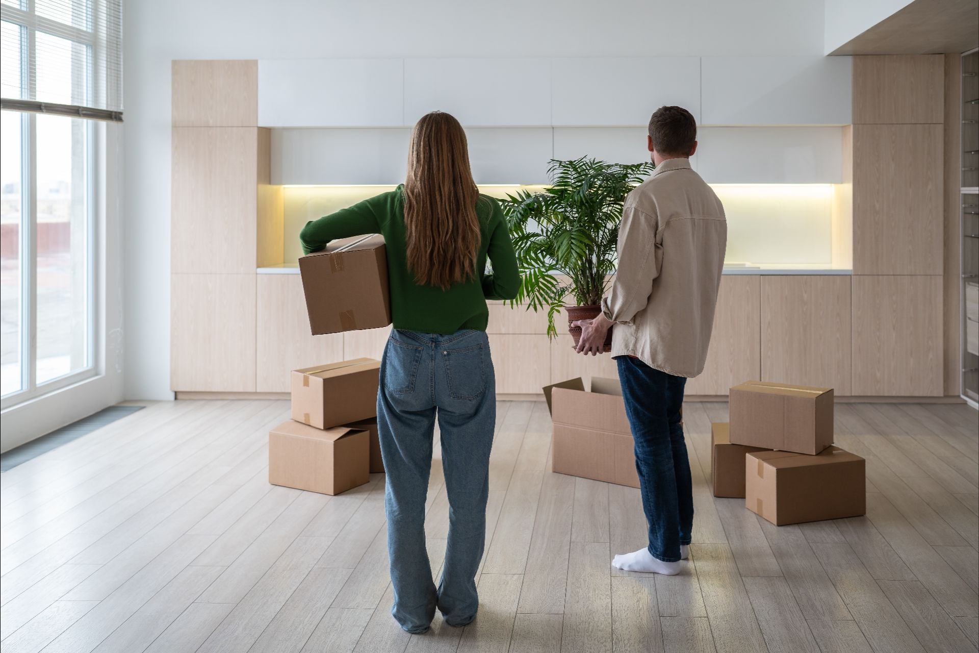 New home owners with boxes and houseplant in hands, rear view. Family couple taking things to another rented light apartment. Housewarming in mortgage apartment. Beginning of happy married life