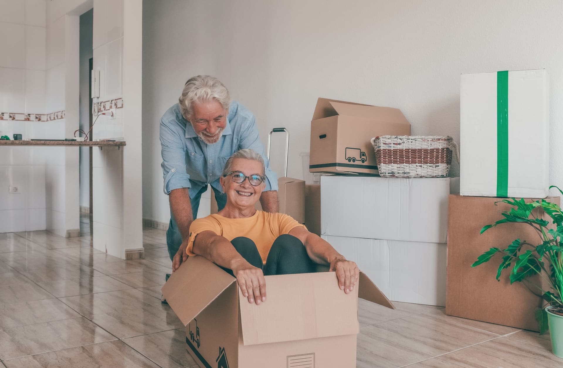 A playful older couple moves into a new home, surrounded by moving boxes in an empty room. A concept of a fresh start.