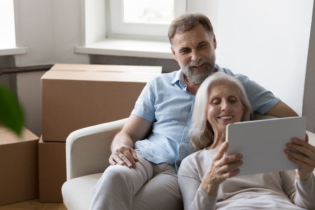 Older couple relax on sofa on move day near boxes with stuff, use tablet buy goods on internet
