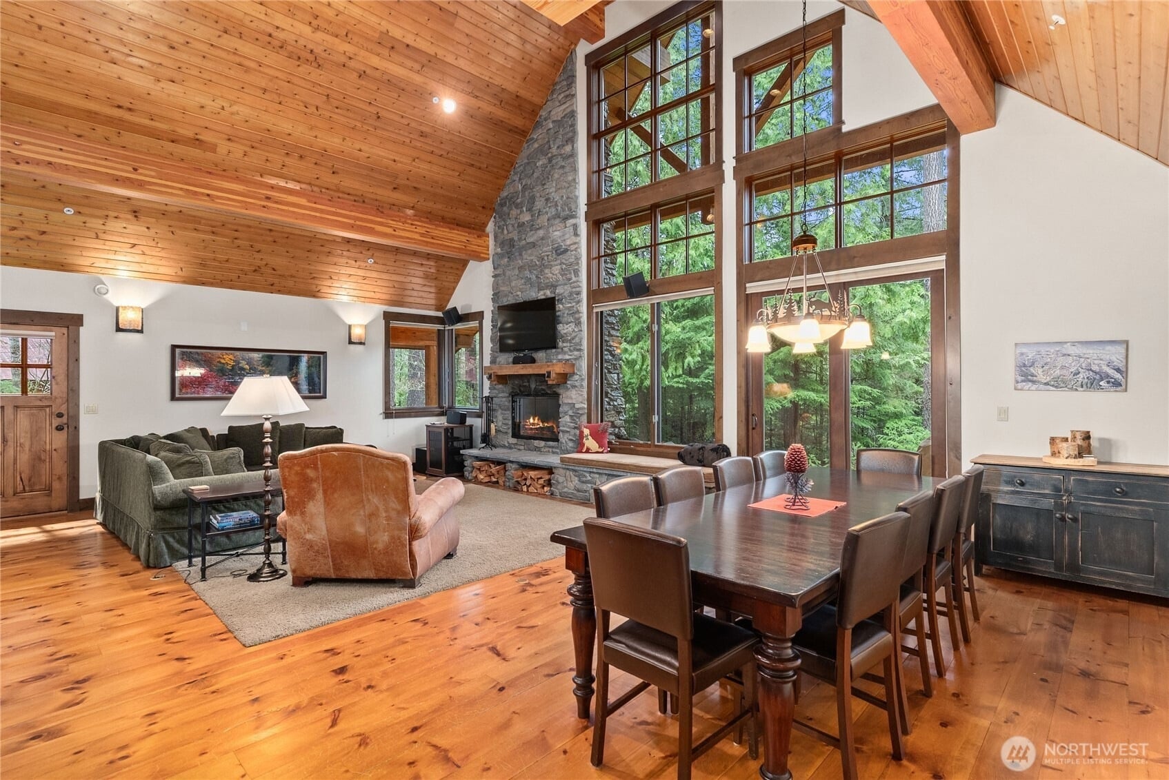 Open-concept living and dining room with vaulted wood ceilings, hardwood floors, a floor-to-ceiling stone fireplace, and large windows overlooking mature evergreen trees.