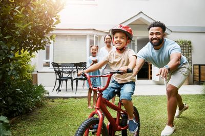 Learning, bicycle and proud dad teaching his young son to ride while wearing a helmet for safety in their family home garden. Active father helping and supporting his child while cycling outside