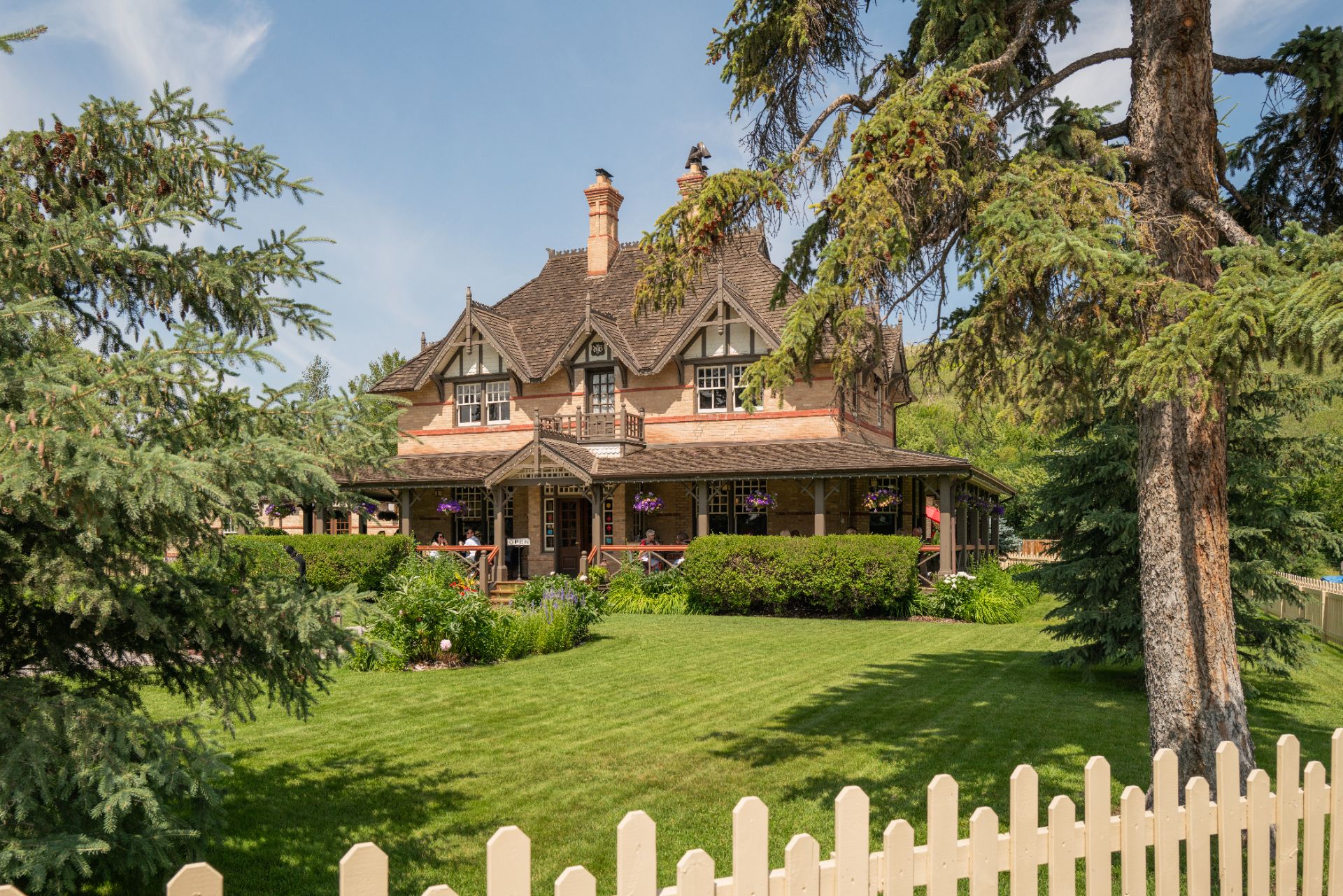 Historic heritage home in Alberta, Canada, surrounded by mature evergreen trees, landscaped gardens, and a manicured lawn on a sunny day.