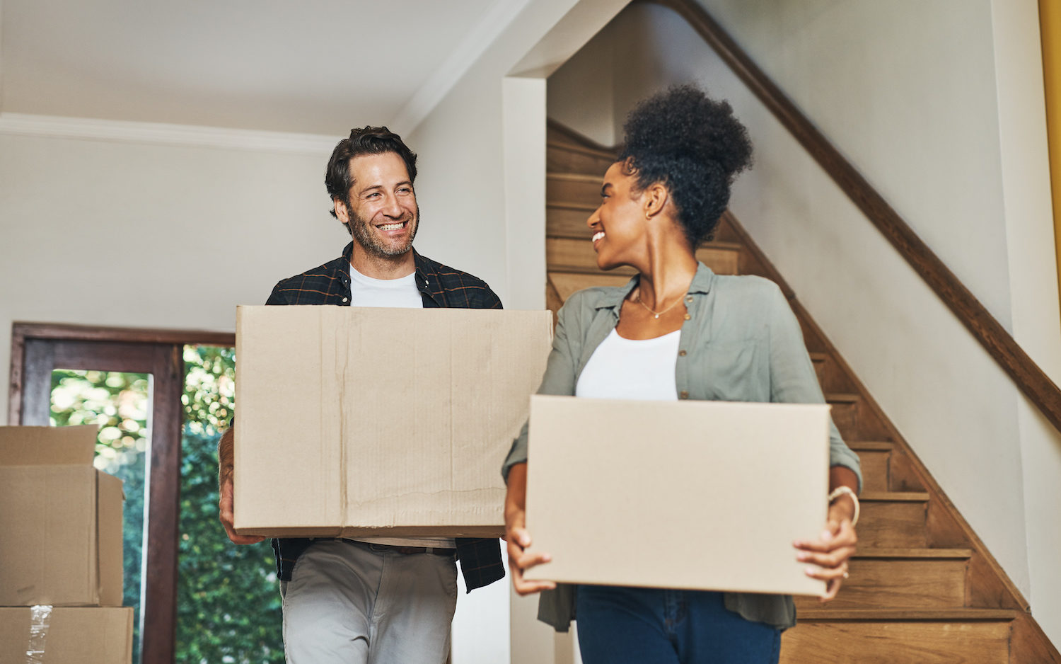 Couple homeowners moving in, carrying boxes and unpacking in new purchased home as real estate investors. Smiling, happy and cheerful interracial man and woman, first time buyers and property owners