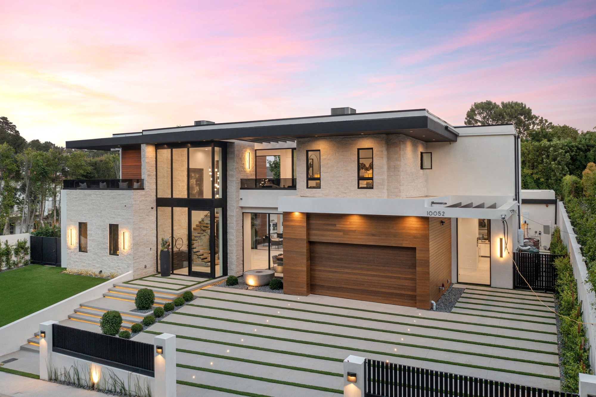 Modern luxury home exterior with large windows, wooden garage door, and well-manicured lawn at sunset