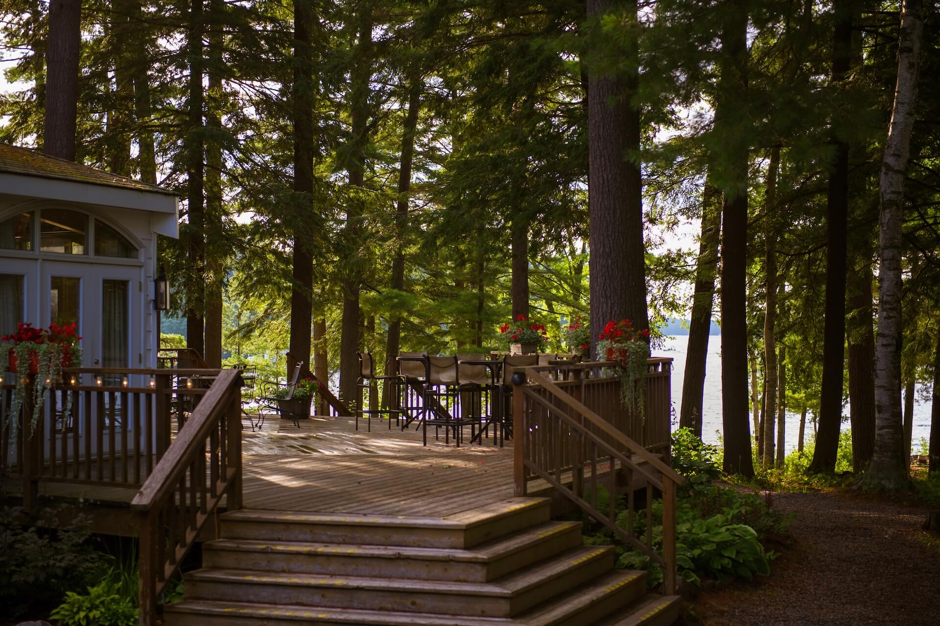 Wooden deck, nestled between trees, with chairs near a lake in Muskoka, Ontario Canada. Red flowers pots are visible.