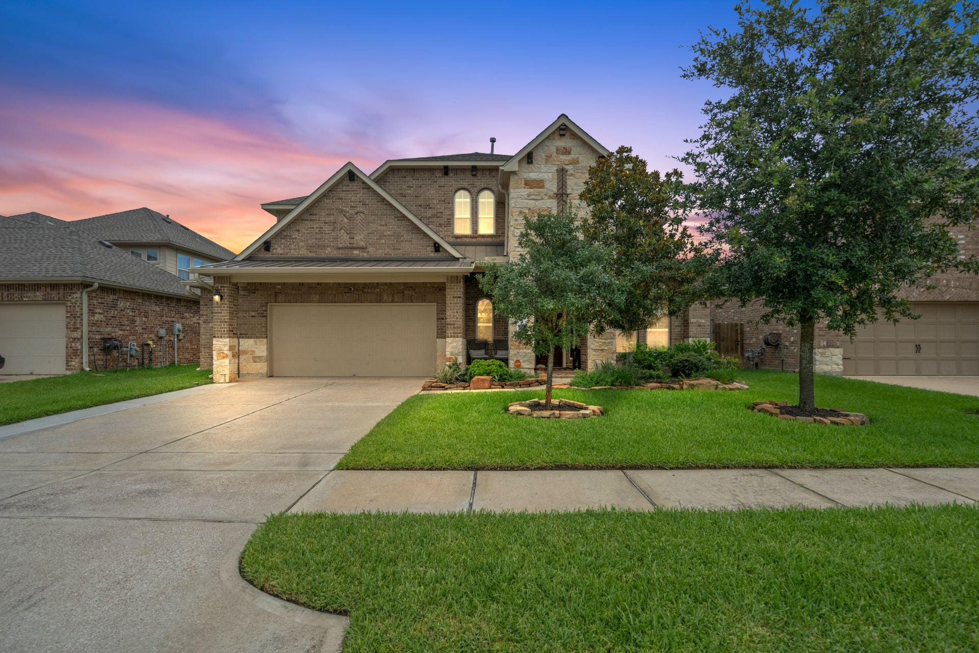 Luxury two-story brick and stone home in Magnolia, Texas, with a three-car garage, manicured lawn, mature trees, and colorful sunset sky in a quiet residential neighborhood.