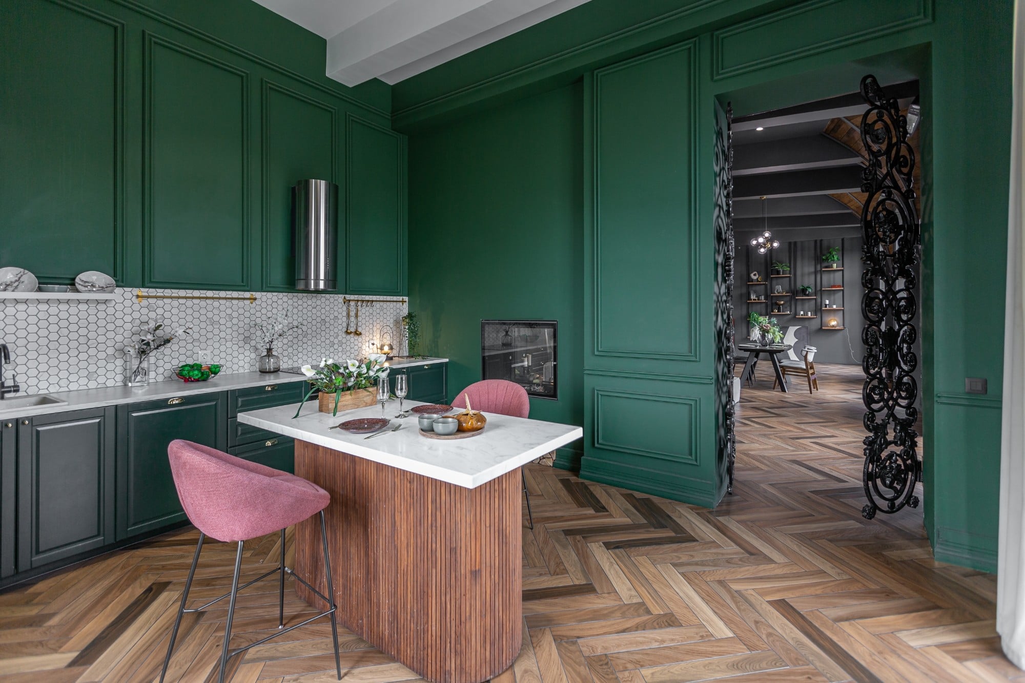 A spacious kitchen showcases a rich green color scheme, featuring a sleek marble island, elegant pink bar stools, and wooden flooring, creating a warm, inviting atmosphere fireplace inside the wall