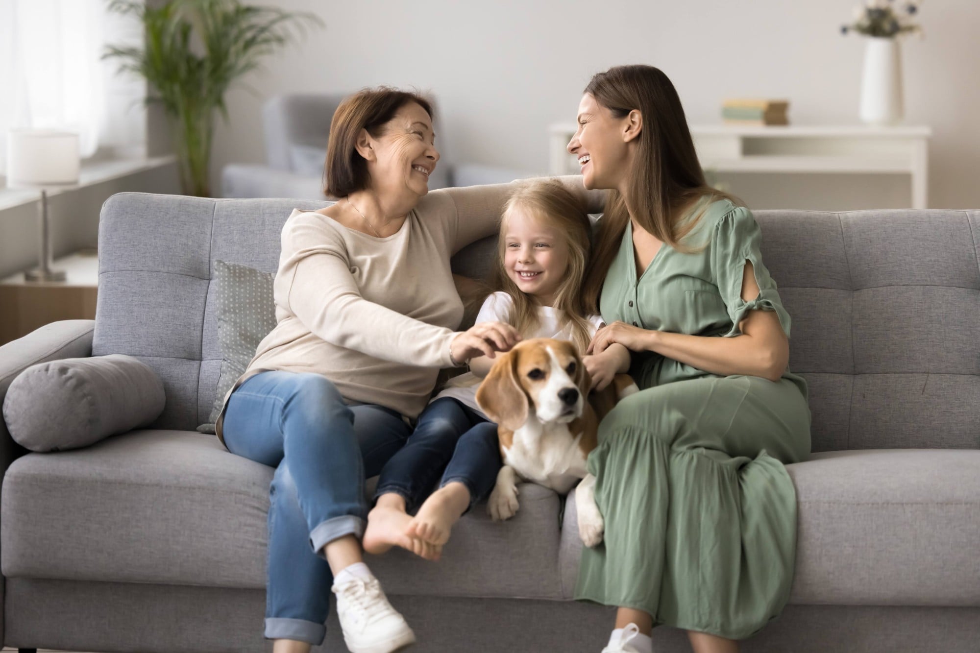 Three generations of women, grandmother, mother, little girl sitting together on sofa with beagle dog, enjoying warm, relaxed moments, exude sense of family bonding, care, and joy, in homey atmosphere (1)