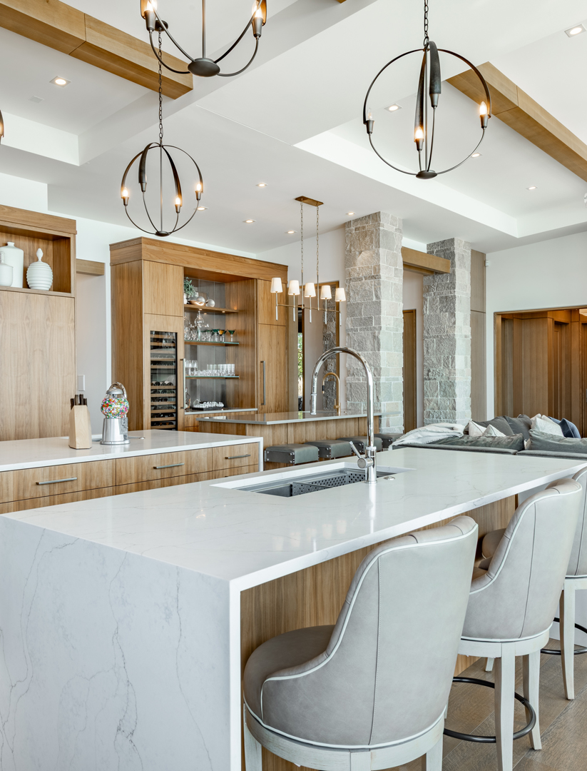 mountain-modern-luxury-kitchen-warm-wood-cabinetry-large-kitchen-island-home-bar-wet-bar-stacked-stone-columns-interior-light-floors-exposed-ceiling-beams-open-concept-living-warmth-rustic-charm