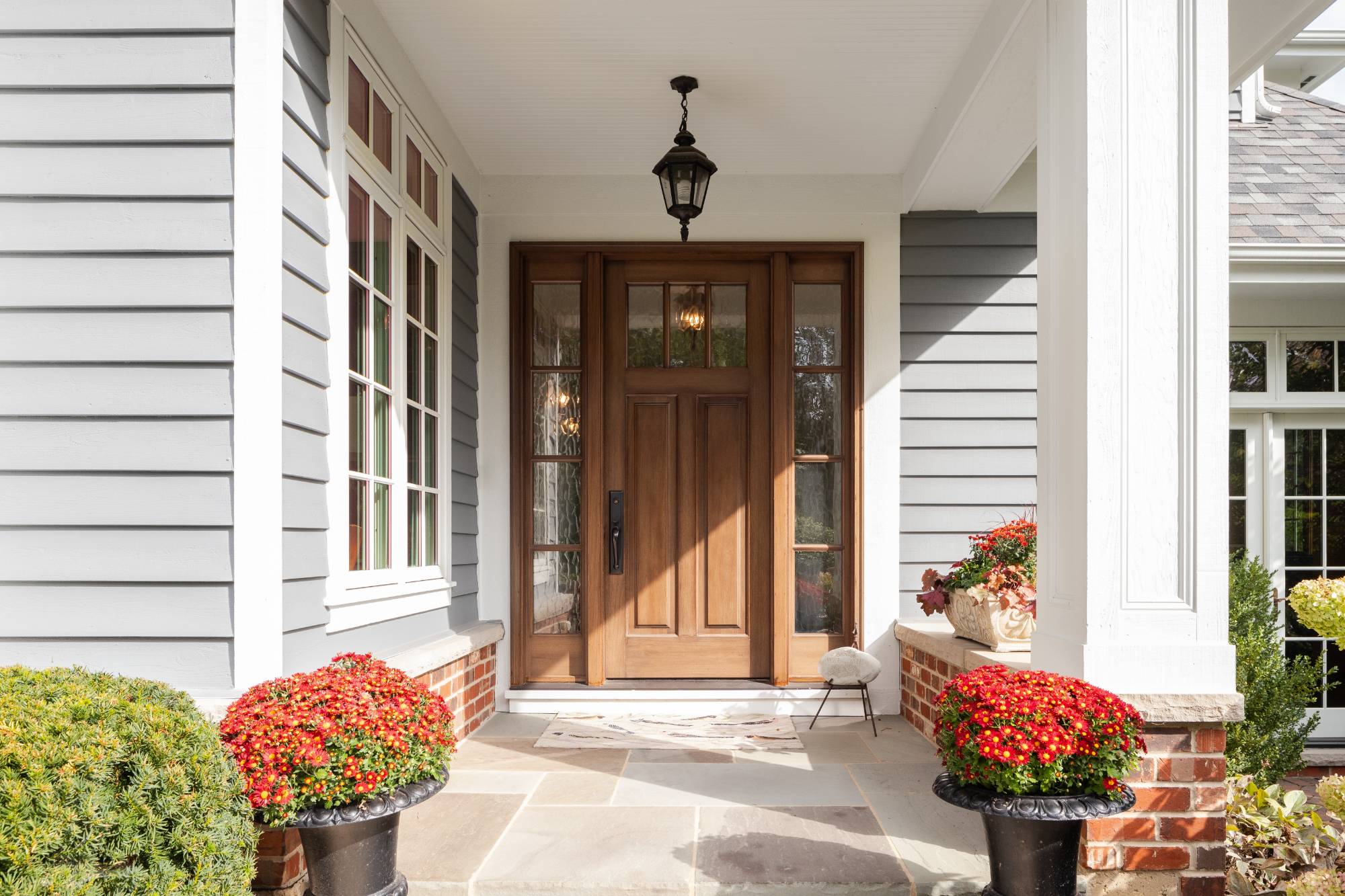 A front door and covered porch detail on a home with grey siding, white trim, red brick, a wooden front door.