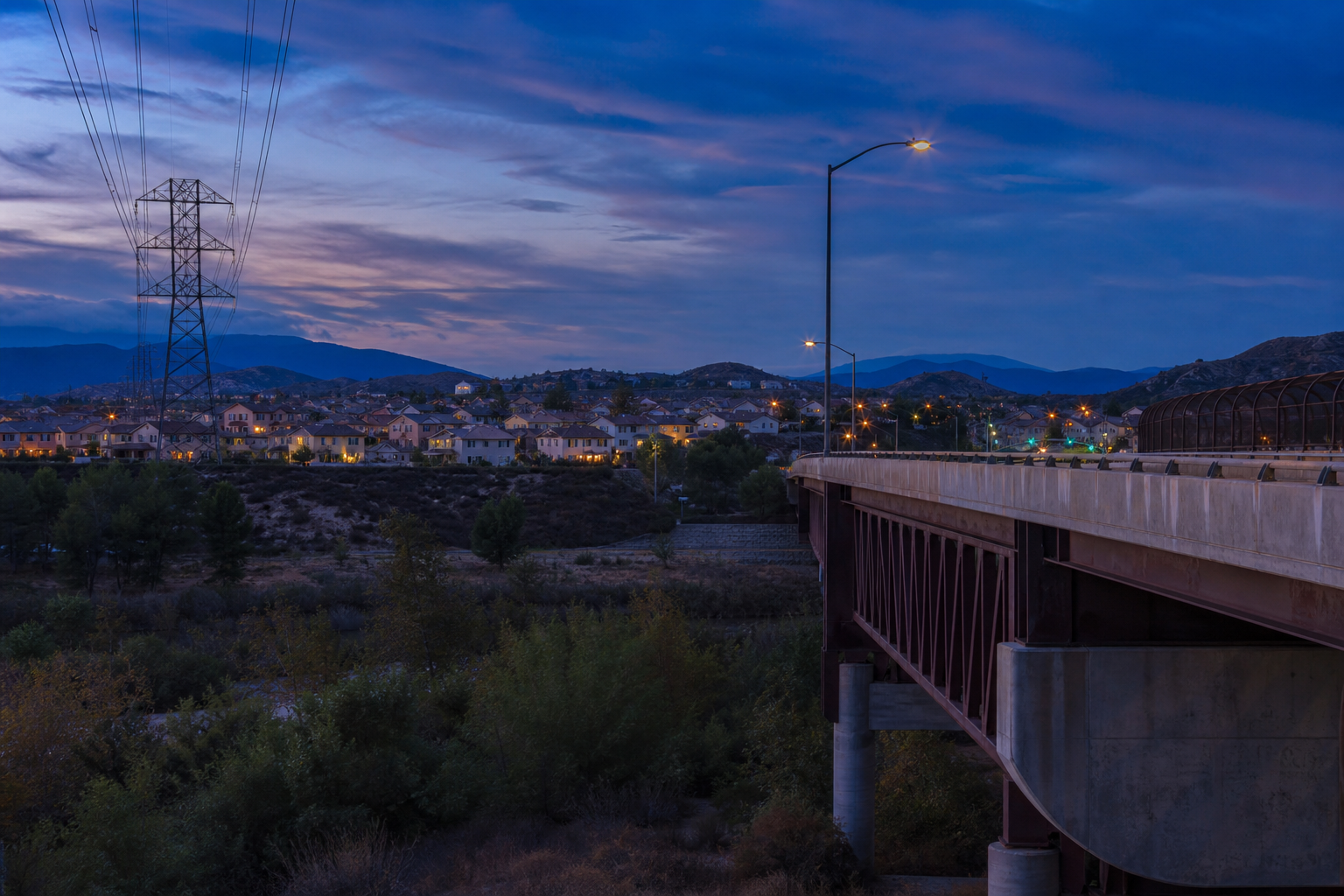 Twilight view of a freeway overpass and power lines with a hillside neighborhood lighting up in the distance in Southwest Riverside County