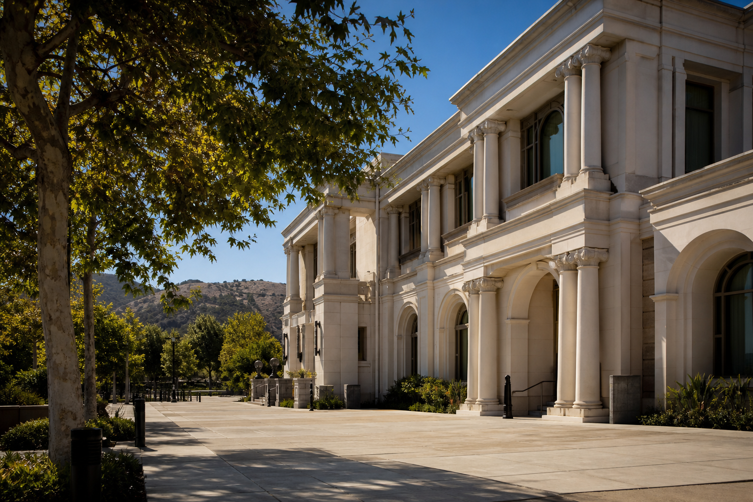 Large cream-colored building with tall columns and arched windows beside a sunlit stone plaza and a shade tree, with hills in the background