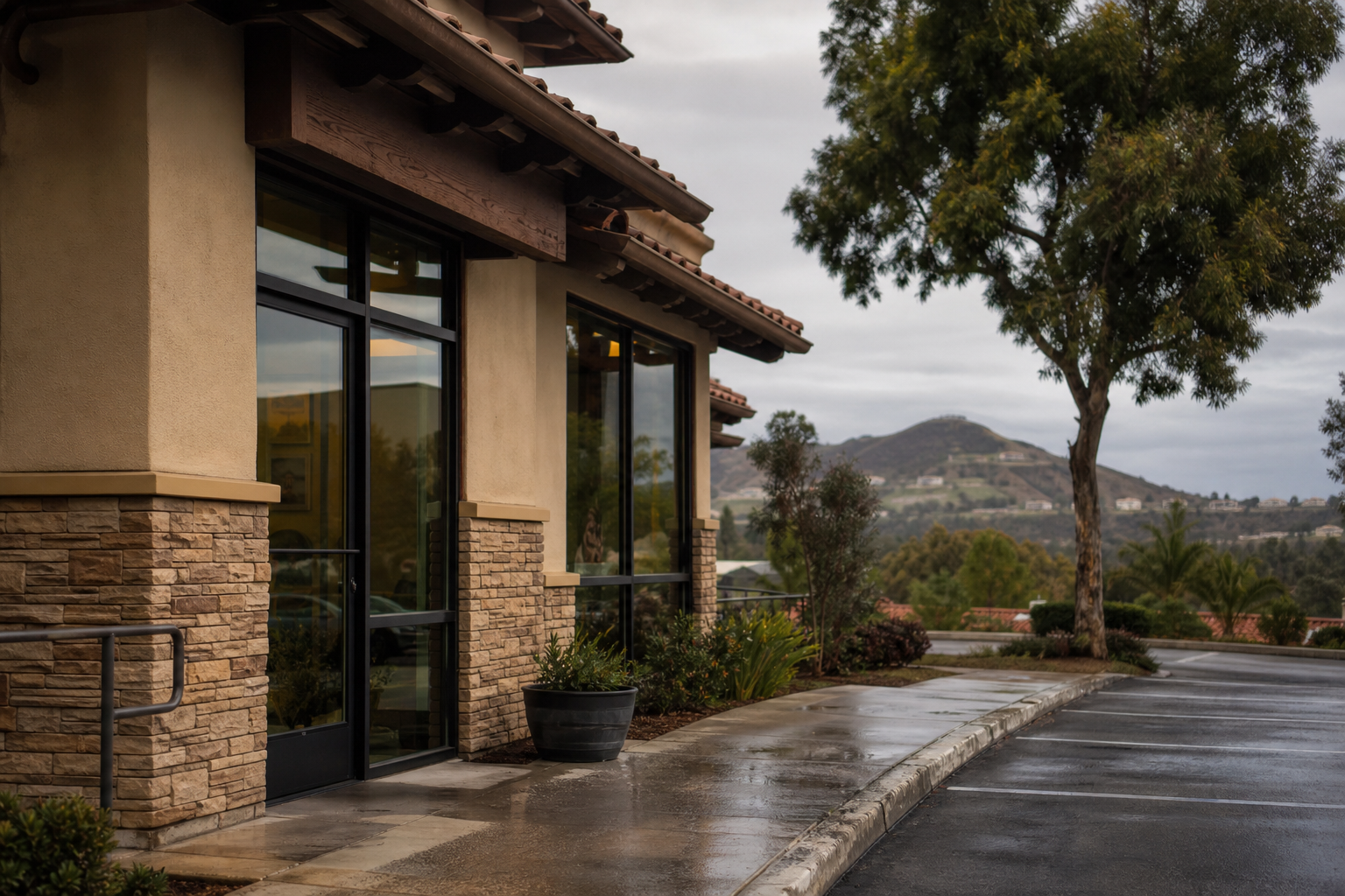 Stucco and stone building with a tile roof and tall windows beside a wet sidewalk, with a tree and rolling hills in the background in Temecula