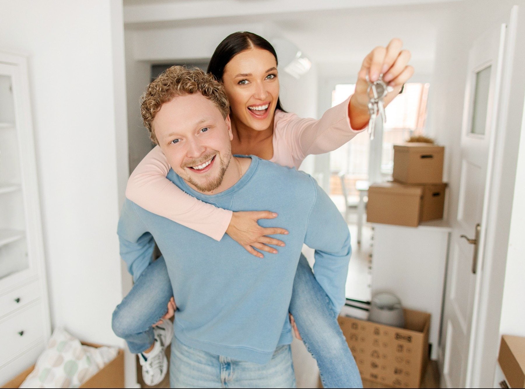 Excited lady getting piggyback ride from her husband and showing house key to camera, posing in new apartment on moving day (1)