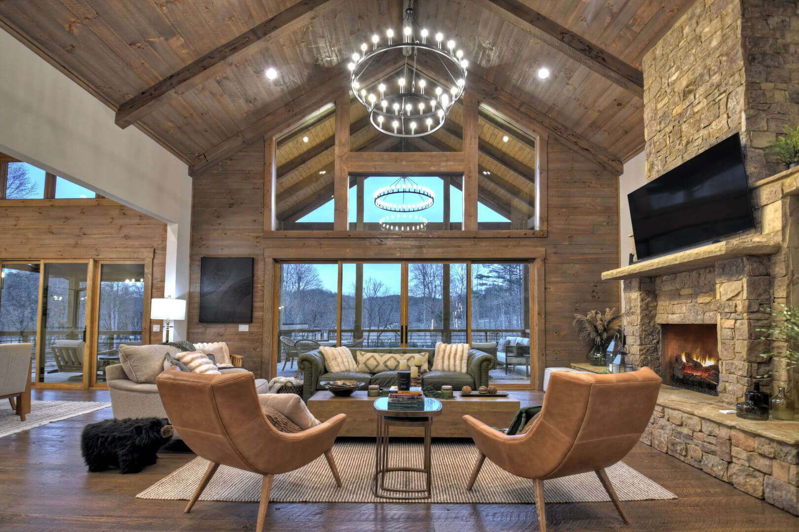 Rustic living room with a stone fireplace, wood paneling, and large windows overlooking nature.