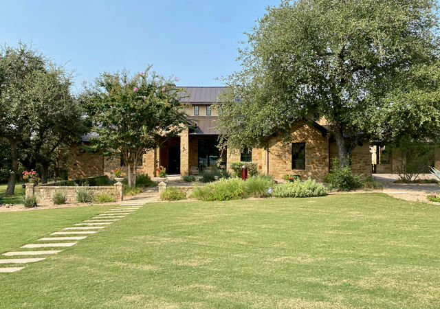 Tan limestone house surrounded by trees and large lawn in front with paver walk