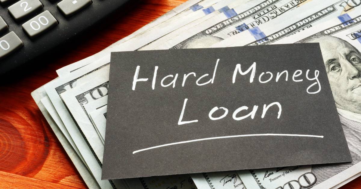 A small, handwritten note reading "HARD MONEY LOAN" resting on a desk on top of a stack of hundred dollar bills.