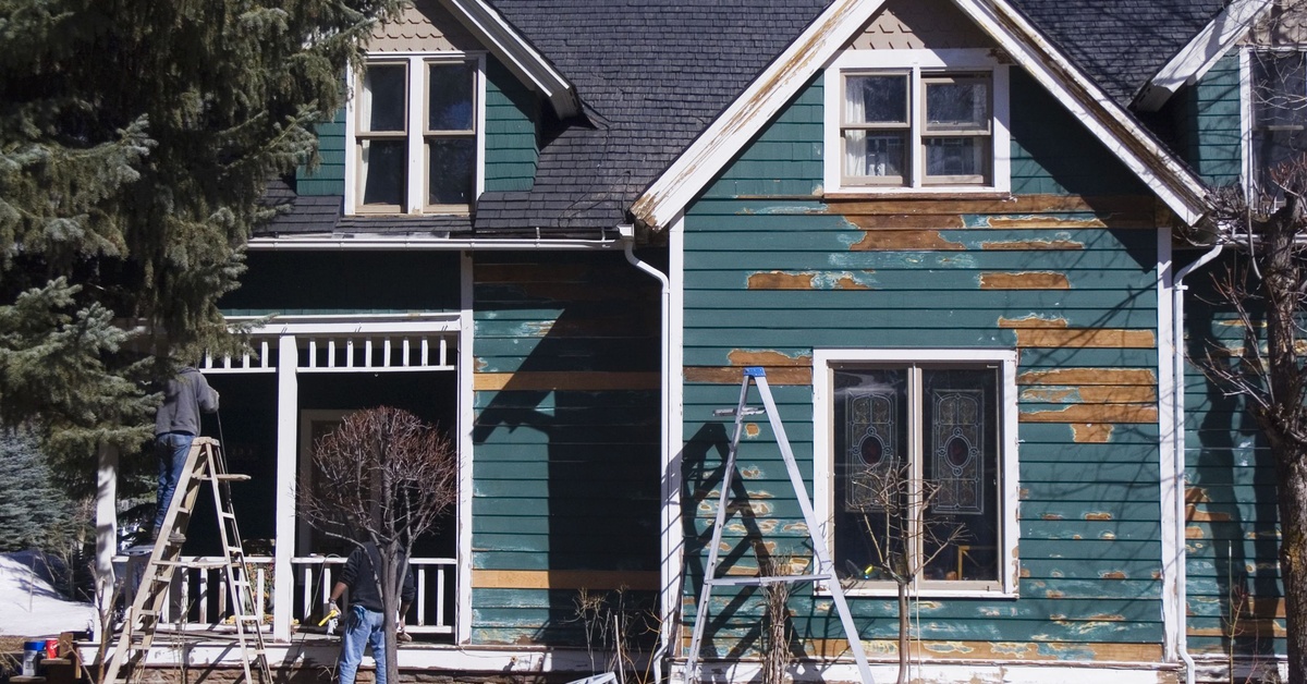 Teal paint peeling from damaged siding on a two-story house with exposed plywood panels and ladders leaning against the wall.
