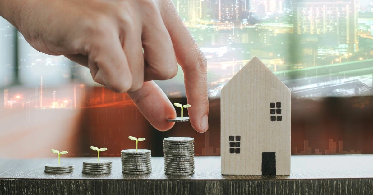 A hand placing a sprout on stacked coins beside a wood house with a city skyline glowing in the background.