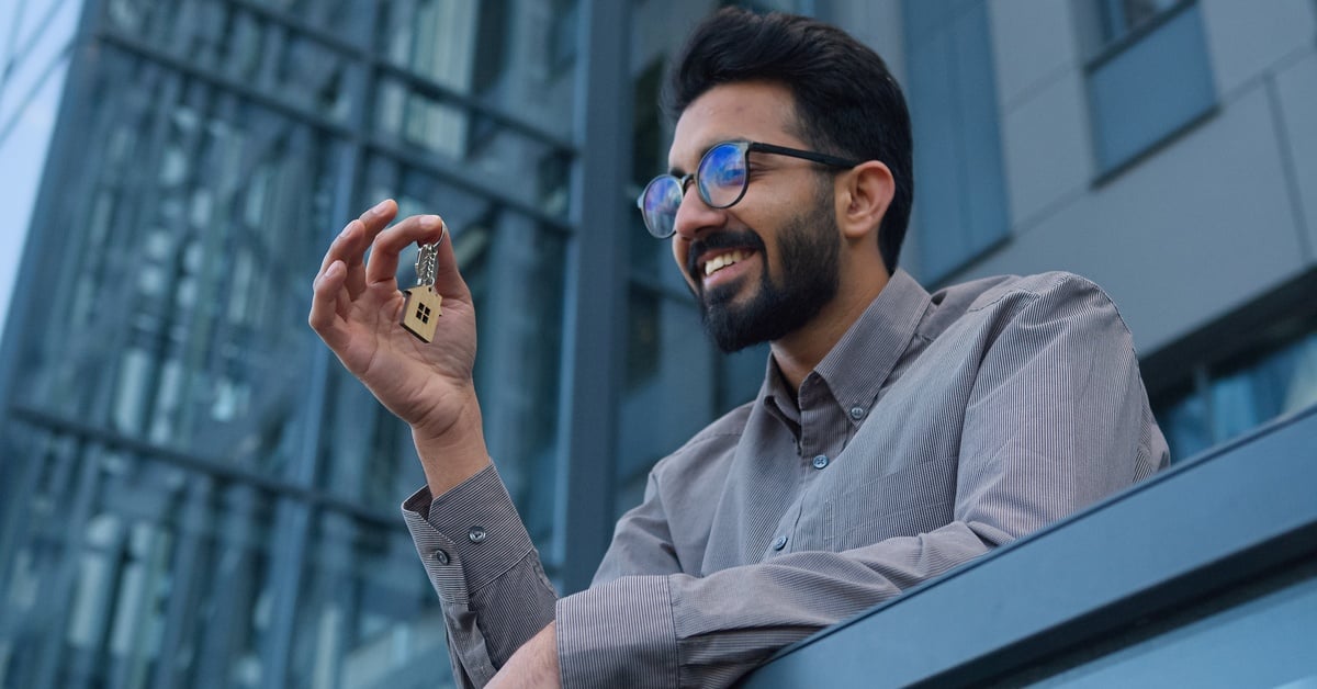 A person standing outside a modern office building smiling while holding keys with a house-shaped keychain.