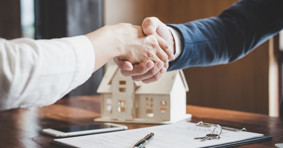 Two people shaking hands over a wooden table holding a contract clipboard, house model, smartphone, pen, and keys.
