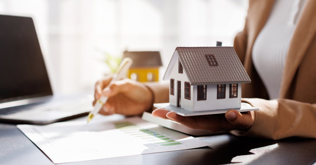 A person in a tan blazer holding a model house beside a paper chart and an open black laptop on a desk.