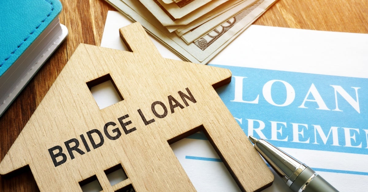A house model with the words "Bridge Loan" sitting on a desk with papers and money nearby.