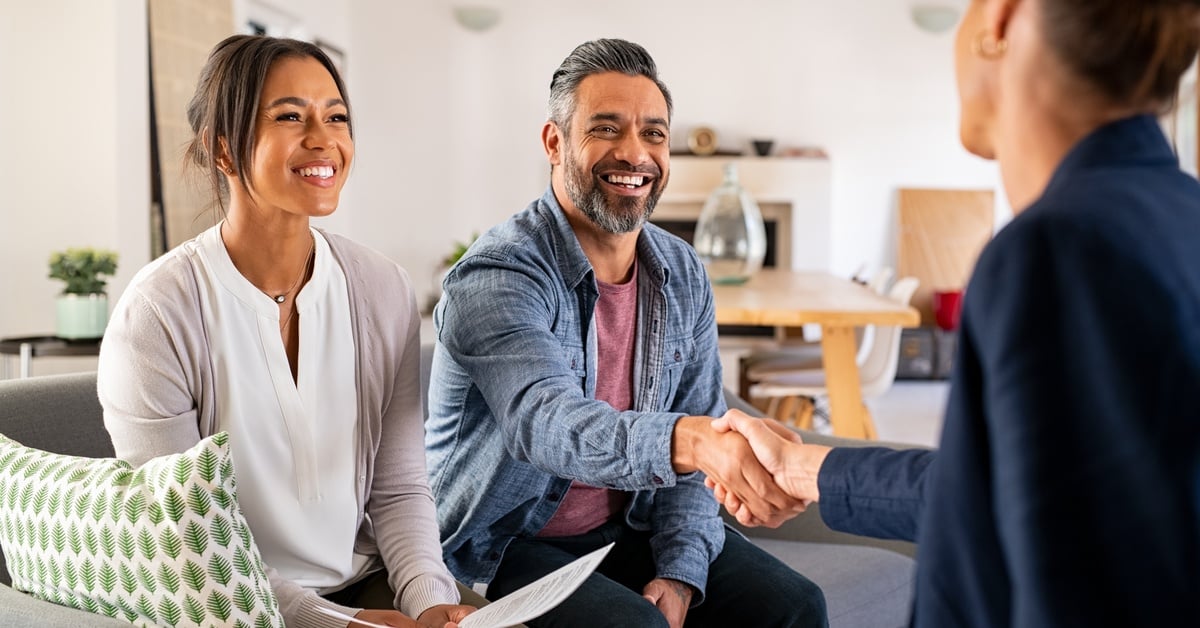 A smiling couple sitting on a couch across from woman. The man is shaking hands with the woman across from him.