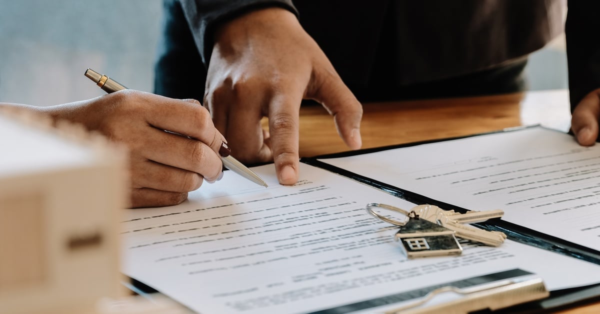 A hand holding a pen while signing a document. Another individual's hand is pointing to a spot on the document.