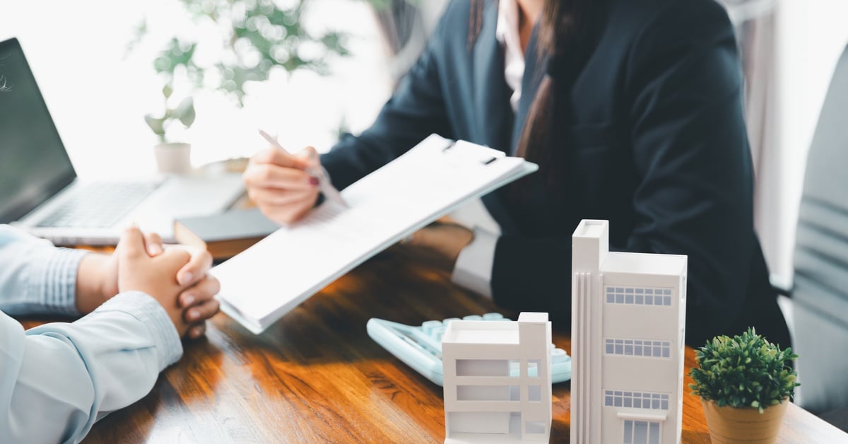 A woman in a dark suit sitting across from another person at a wooden desk. She's holding a pen and paper, indicating a specific spot on the paper to the person across from her
