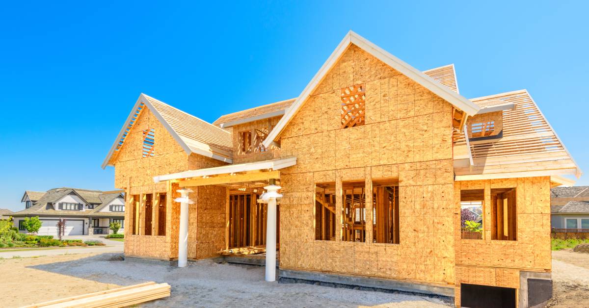 A wood frame house under construction with visible exposed plywood, roof trusses, and white column porch supports.