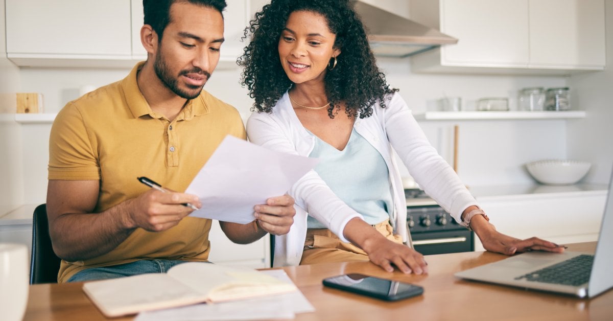 A man and woman in a modern white kitchen with papers, a notebook, phone, and laptop on a wood table.