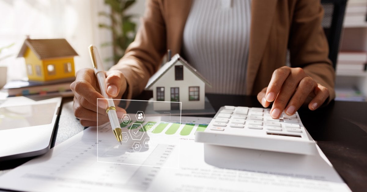 A person in blazer using calculator at office desk with a house model, tablet, documents, and energy chart on it.