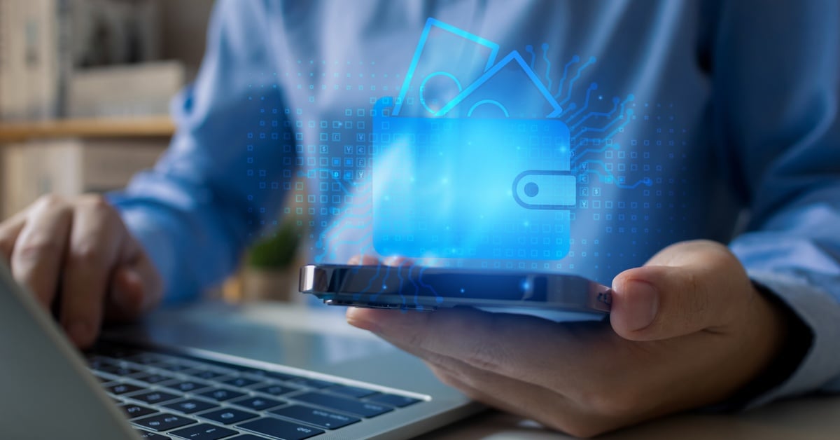 A man in a blue shirt holding a smartphone with a glowing blue wallet hologram above it while using a laptop at a wooden desk.