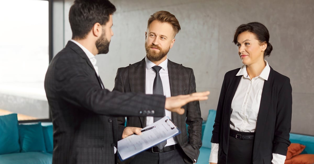 Three businesspeople in formal attire discussing paperwork while standing inside a modern office.