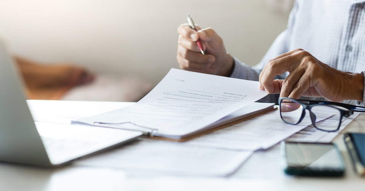 A person reviewing documents at a desk with a pen in their hand. Eyeglasses and a laptop are beside the papers.